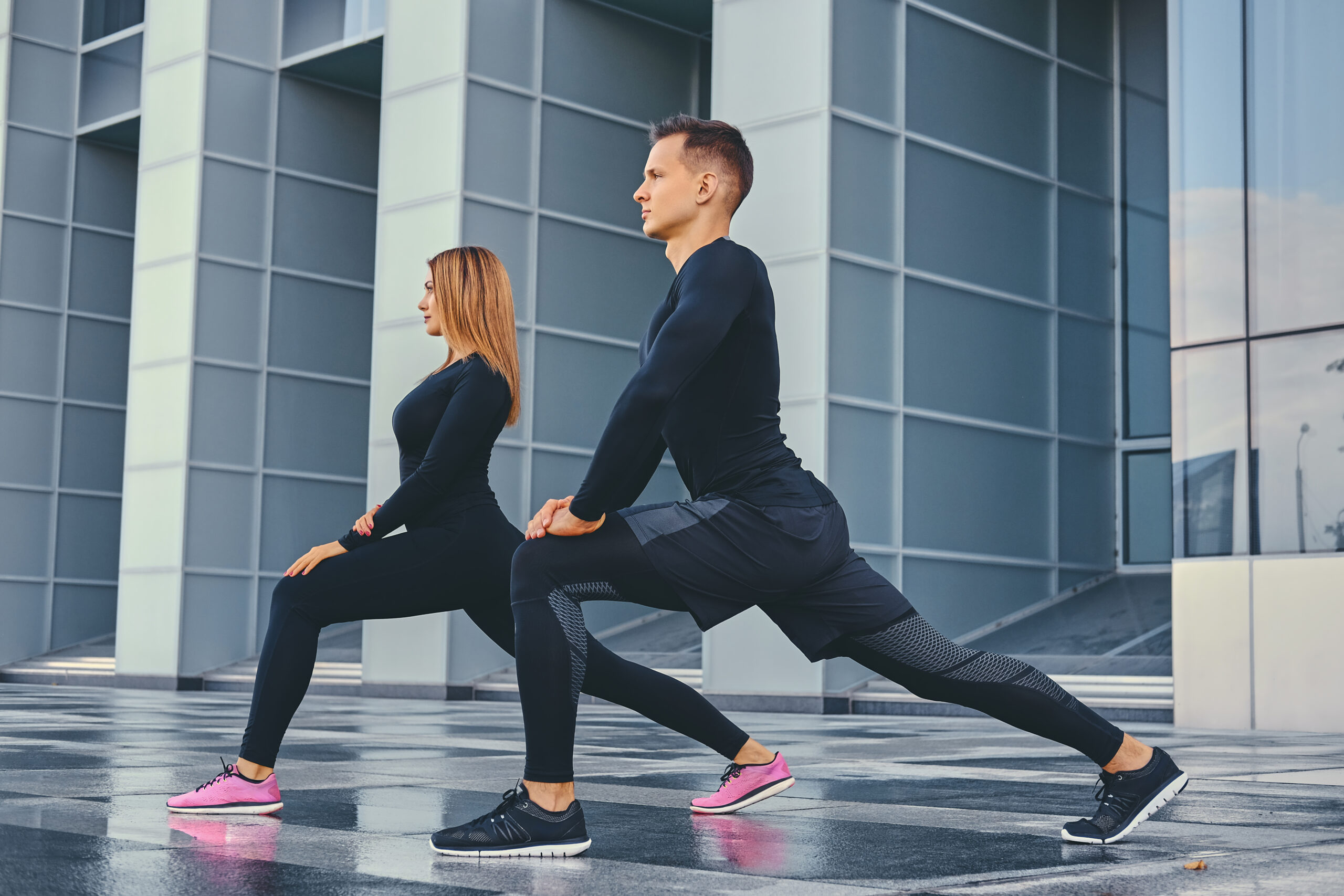 The athletic fitness couple is stretching over modern building background. Full body image.